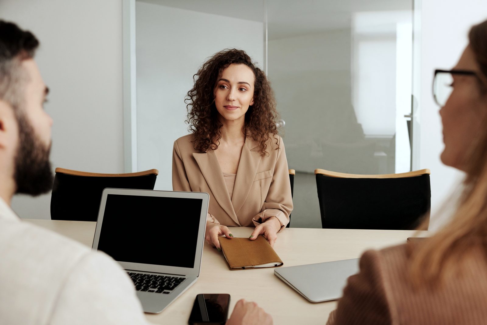 A candidate in a camel blazer, seated at the table, listens attentively to two interviewers in a bright office