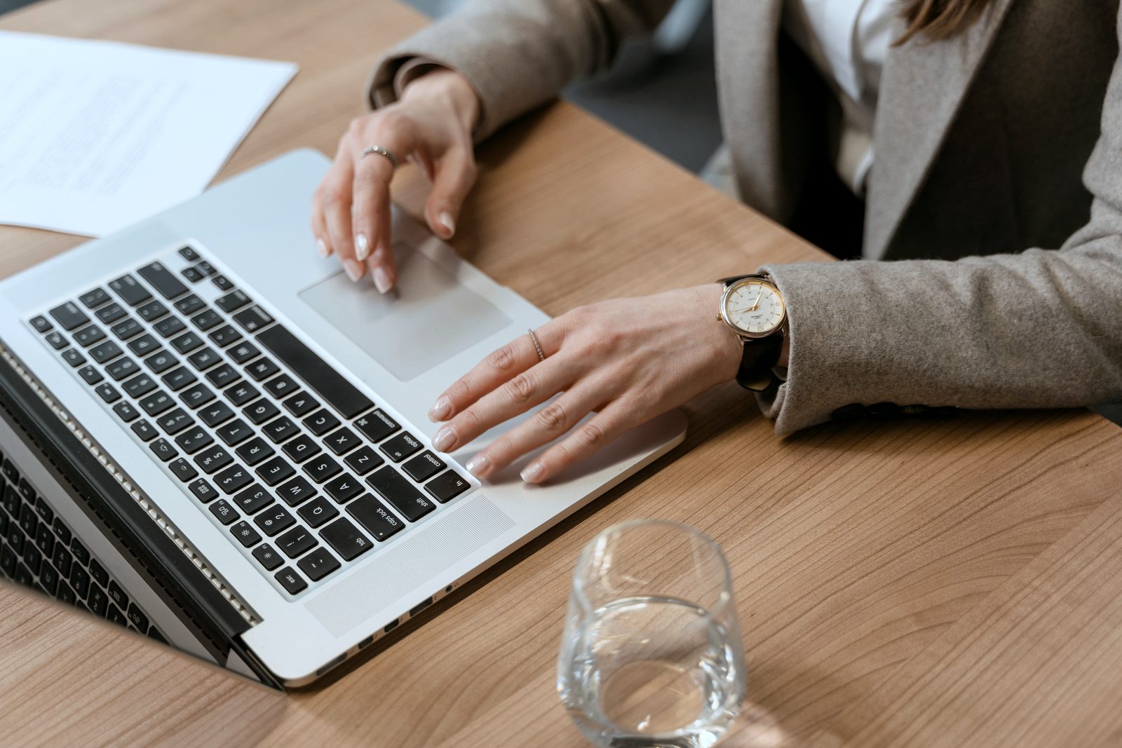 Hands on a laptop on a light wooden table, next to documents and a glass of water